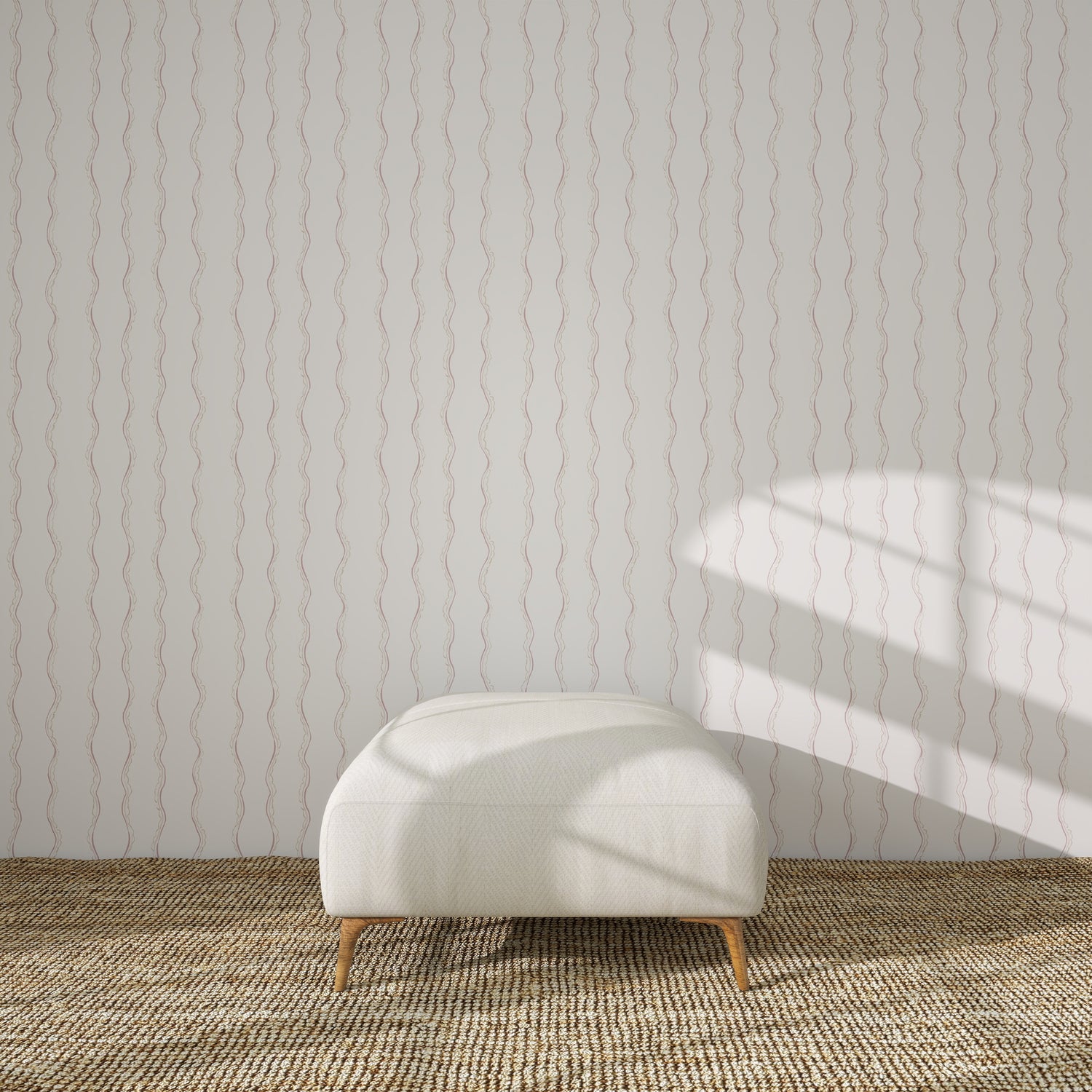 A white upholstered bench with wooden legs sits on a woven rug, against a white wall adorned with Ribbon Stripe Wallpaper in Rose. Sunlight casts gentle shadows, creating a serene scene perfect for elegant children&