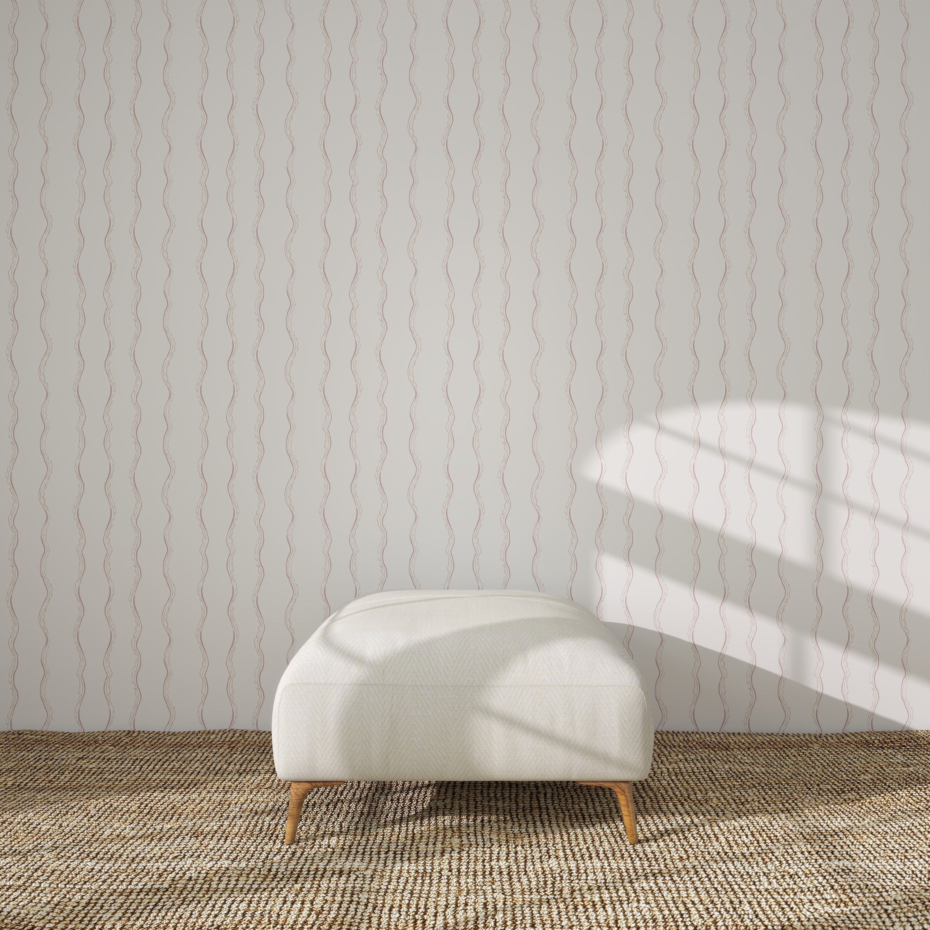 A white upholstered bench with wooden legs sits on a woven rug, against a white wall adorned with Ribbon Stripe Wallpaper in Rose. Sunlight casts gentle shadows, creating a serene scene perfect for elegant children&