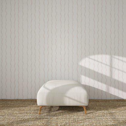 A white upholstered bench with wooden legs sits on a woven rug, against a white wall adorned with Ribbon Stripe Wallpaper in Rose. Sunlight casts gentle shadows, creating a serene scene perfect for elegant children&
