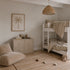 A neutral-toned kids’ bedroom features bunk beds, a woven pendant lamp, a palm tree art piece, a cozy floor pillow, a rug, two toy cars, and a dresser adorned with plush toys. The subtle coastal nursery decor theme is beautifully highlighted by the gentle touch of the Scallop Shell Mural in Mustard. Soft elements like nursery wallpaper and seaside accents create a calm and cozy atmosphere in the room.