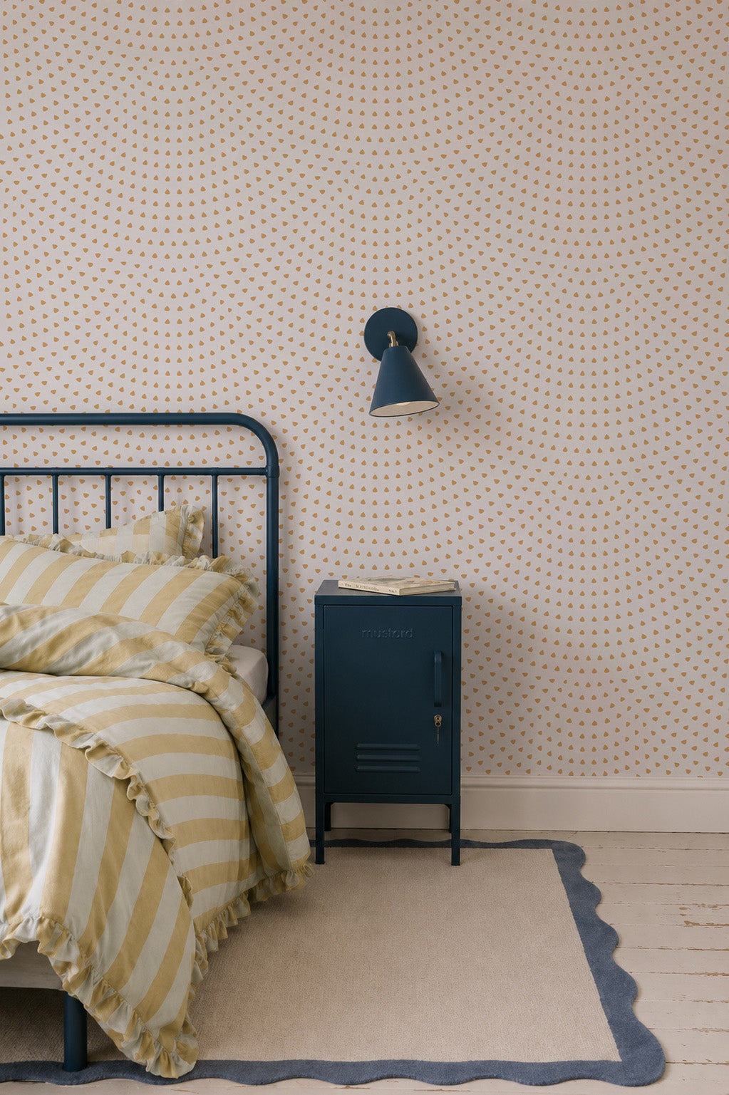 A cozy bedroom corner featuring a striped yellow and white bed, a blue metal nightstand, and a wall-mounted blue lamp is brought to life by the Scallop Shell Mural in Mustard, creating charming coastal nursery decor. A beige rug with a blue wavy border covers the wooden floor.