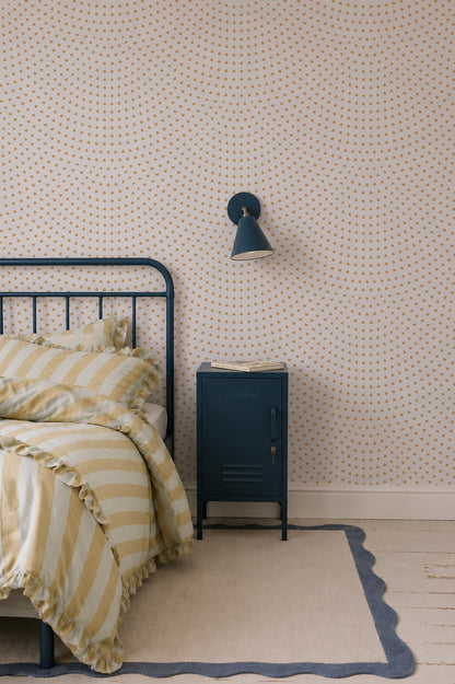 A cozy bedroom corner featuring a striped yellow and white bed, a blue metal nightstand, and a wall-mounted blue lamp is brought to life by the Scallop Shell Mural in Mustard, creating charming coastal nursery decor. A beige rug with a blue wavy border covers the wooden floor.