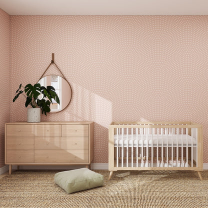 A nursery featuring Scallop Shell Wallpaper Repeat Pattern | Rose, a wooden crib, matching dresser with potted plant and round mirror, woven rug, and green cushion—perfect as a cozy playroom.