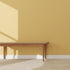 A wooden bench rests against a wall covered in Scallop Shell Wallpaper Yellow | Sample. Sunlight casts shadows, while a chevron rug on the wooden floor creates an inviting space inspired by online imagery.