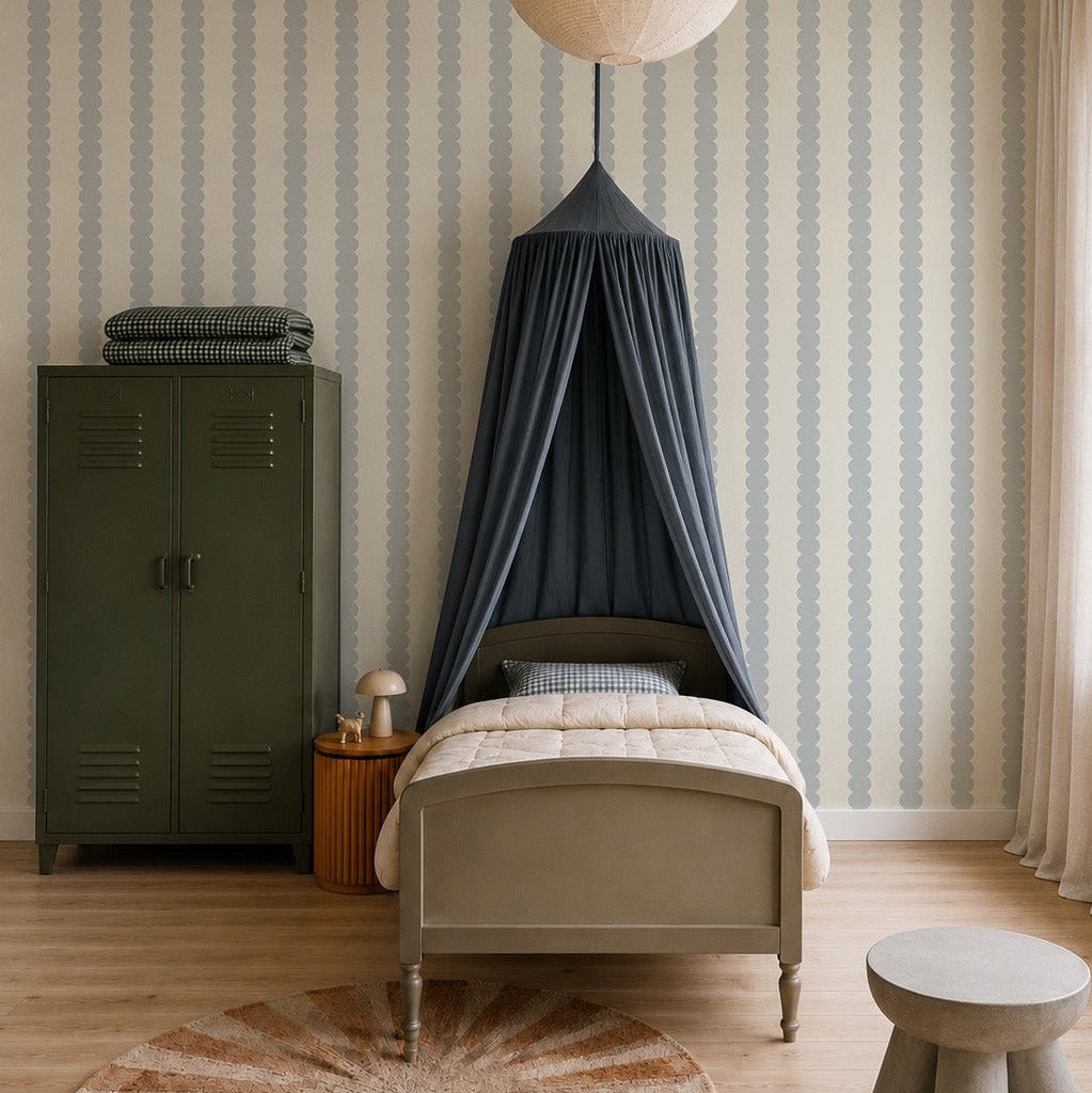 A cozy kids’ bedroom with a gray bed under a dark blue canopy, a green locker wardrobe, a small round table and stool, and a large paper lantern. The walls feature Scallop Stripe Wallpaper, Blue for added charm.