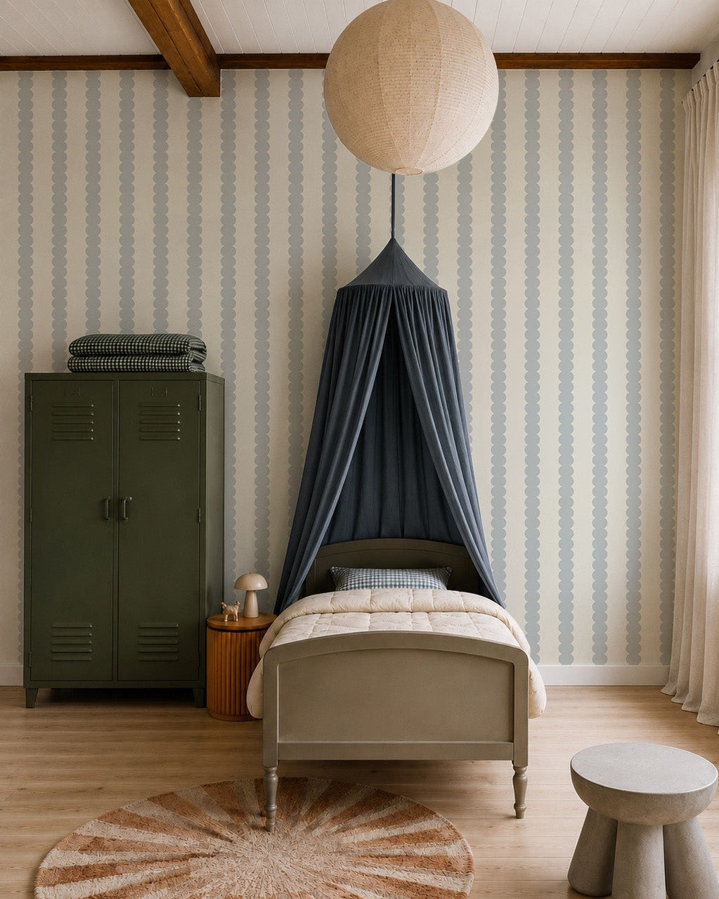 A cozy kids’ bedroom with a gray bed under a dark blue canopy, a green locker wardrobe, a small round table and stool, and a large paper lantern. The walls feature Scallop Stripe Wallpaper, Blue for added charm.