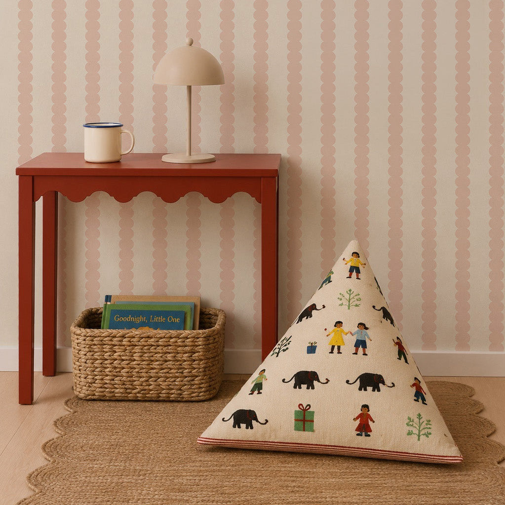 A red table with a lamp and mug sits against blush Scallop Stripe Wallpaper. Below, a woven basket holds two books, and a triangular cushion with colorful animals rests on a scalloped rug.
