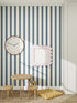 A children’s playroom with a small wooden table and stools, a yellow floor cushion, a large wall clock, a framed picture, and Scallop Stripe Wallpaper Deep Blue—ideal for creating a cozy nursery atmosphere.