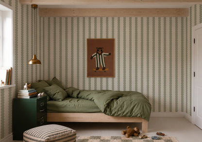 A cozy bedroom featuring a green bed, Scallop Stripe Wallpaper in Green, a bear illustration on the wall, a gold lamp, green cabinet, books, a striped ottoman, and toys on the floor. Soft natural light comes from a window on the left.