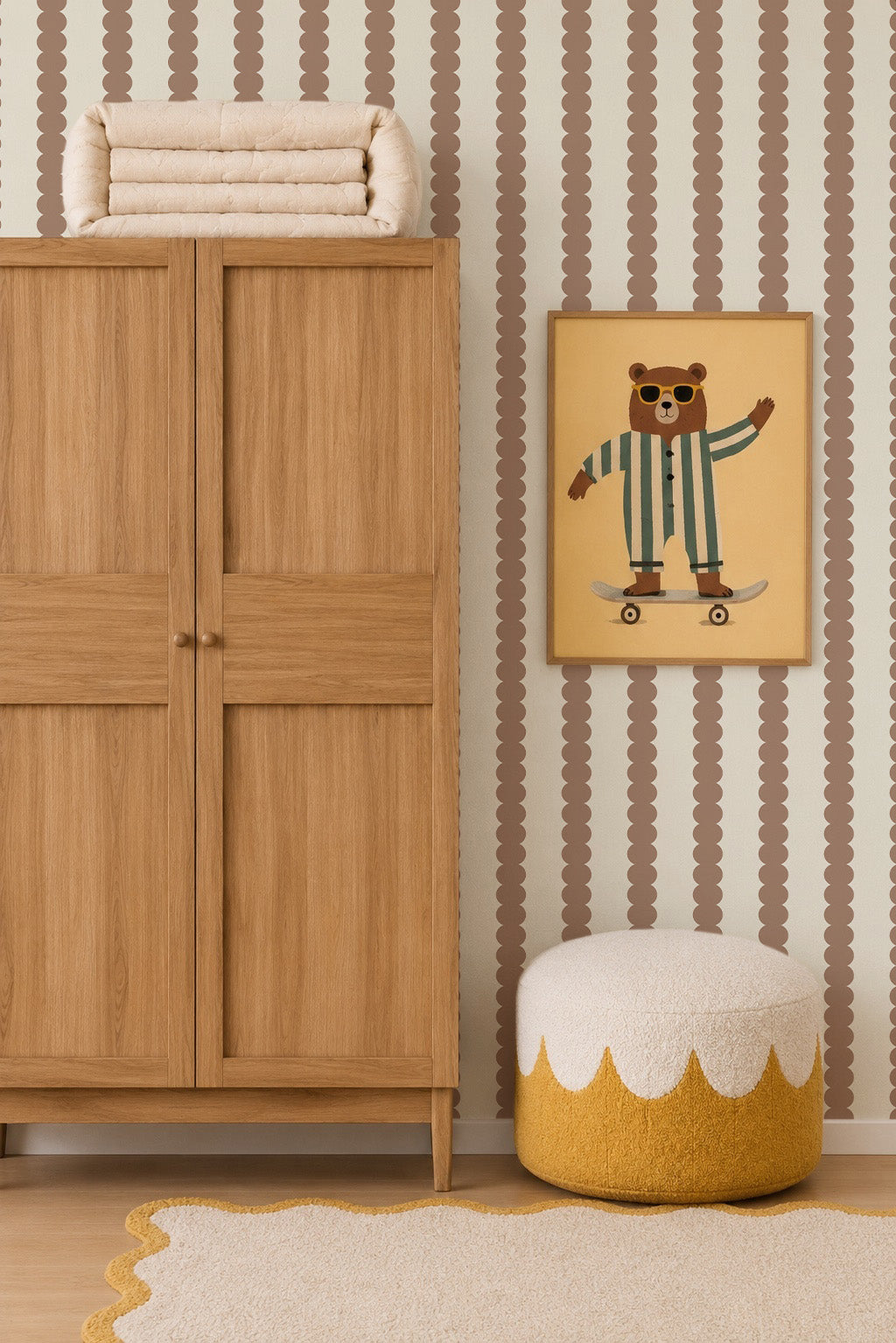 A cozy kids’ room features a wooden wardrobe, white and yellow pouf, beige scalloped rug, and Scallop Stripe Wallpaper in Latte. A framed bear-on-skateboard picture hangs on the wall, with blankets stacked atop the wardrobe.