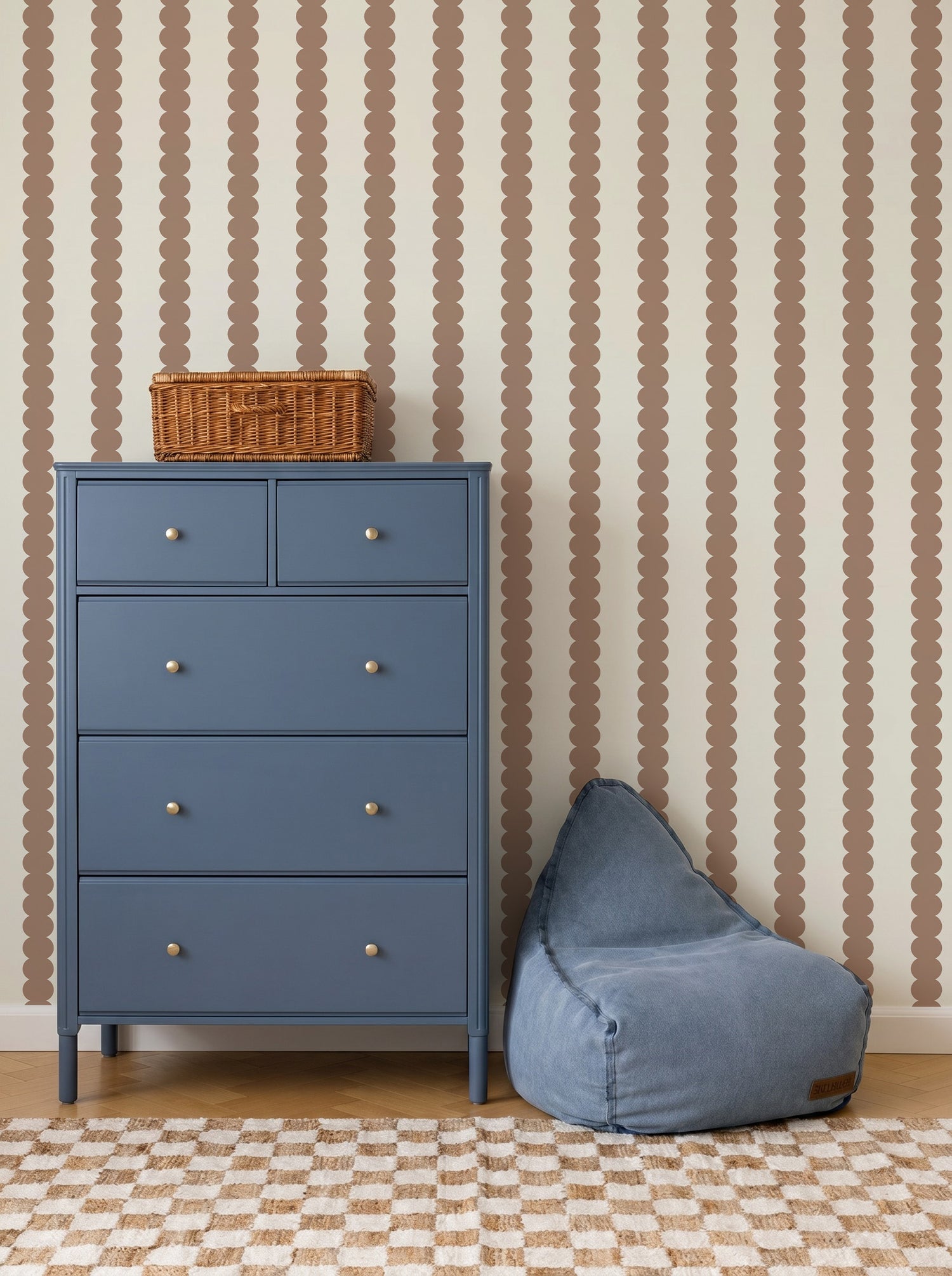 A blue dresser with five drawers stands next to a blue beanbag chair. Behind them, the beige wall is decorated with Scallop Stripe Wallpaper in Latte, featuring a brown scalloped stripe pattern. A woven basket sits on top of the dresser.