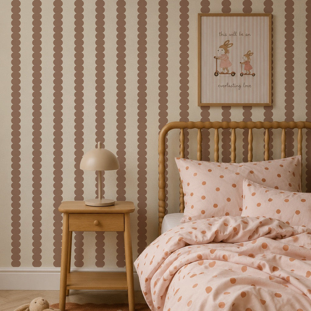 A cozy bedroom features a wooden bed with polka dot bedding, a matching nightstand, and a lamp. The kids’ room walls are adorned with the Scallop Stripe Wallpaper in Latte, adding warm and playful vertical brown scalloped stripes. Above the bed, framed art depicting animals reads “this will be an everlasting love.”.