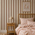 A cozy bedroom features a wooden bed with polka dot bedding, a matching nightstand, and a lamp. The kids’ room walls are adorned with the Scallop Stripe Wallpaper in Latte, adding warm and playful vertical brown scalloped stripes. Above the bed, framed art depicting animals reads “this will be an everlasting love.”.