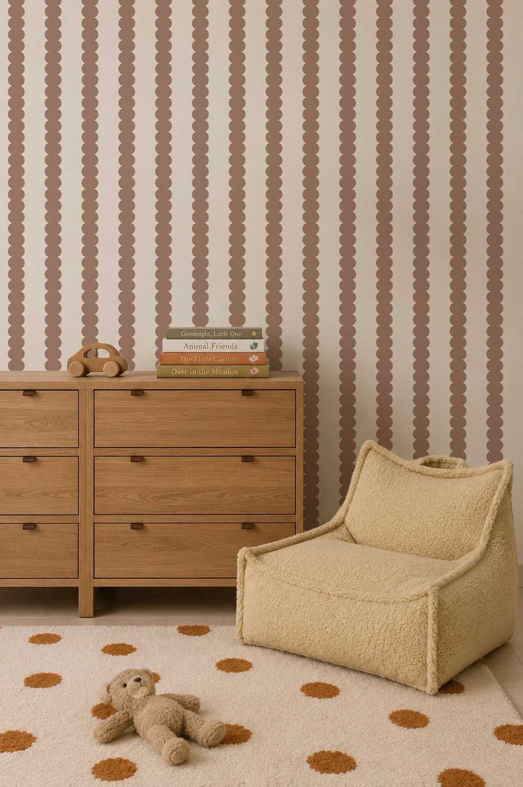 A cozy room with a wooden dresser, stacked children’s books, a toy phone, a plush chair, a polka-dot rug, and a stuffed bear. The wall features Scallop Stripe Wallpaper in Latte—an ideal choice for stylish nursery wallpaper or playful kids’ room wallpaper.