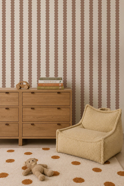 A cozy room with a wooden dresser, stacked children’s books, a toy phone, a plush chair, a polka-dot rug, and a stuffed bear. The wall features Scallop Stripe Wallpaper in Latte—an ideal choice for stylish nursery wallpaper or playful kids’ room wallpaper.