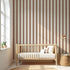 A minimal nursery features a wooden crib, neutral bedding, a basket, and wall lamp. Arched windows highlight the colour and quality of the Scallop Stripe Wallpaper Sample in Mocha and a round jute rug on the light floor.