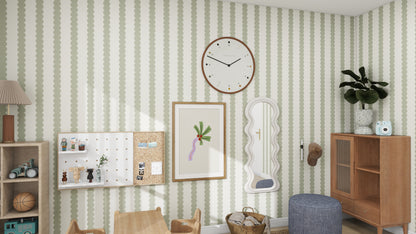 A childrens room features munks and mes Scallop Stripe Wallpaper Green with playful scallop patterns, a large wall clock, art pieces, wooden table and chairs, a cabinet with toys and a plant on top, shelves filled with toys, and a blue pouffe on the floor.