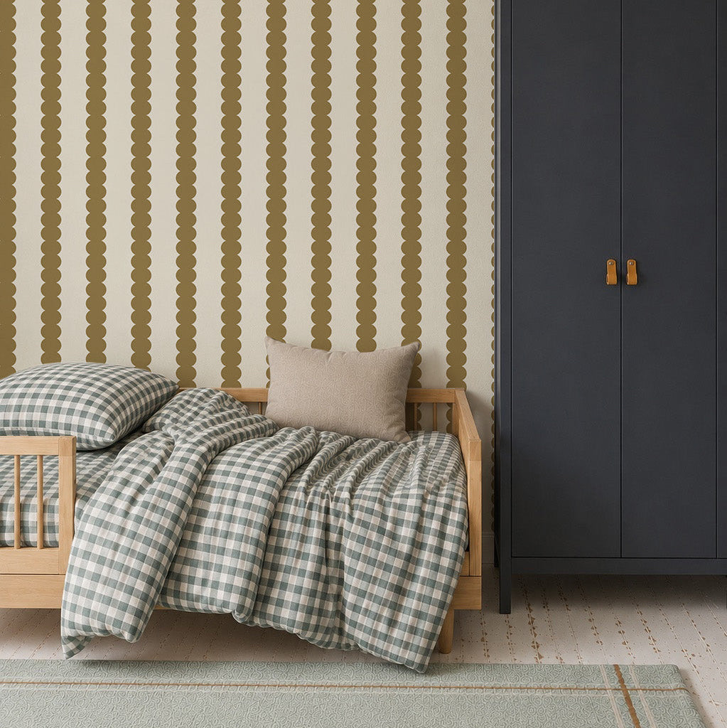 A cozy children’s room features a small wooden bed, checkered bedding, a beige pillow, a tall dark blue wardrobe, and Scallop Stripe Wallpaper in Moss. A large blue pendant lamp hangs overhead.