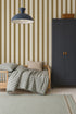 A cozy children’s room features a small wooden bed, checkered bedding, a beige pillow, a tall dark blue wardrobe, and Scallop Stripe Wallpaper in Moss. A large blue pendant lamp hangs overhead.
