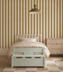 A cozy bedroom with Scallop Stripe Wallpaper in moss, a cream bed with textured bedding, a pastel green storage bench, wooden dresser, geometric rug, and a gold pendant lamp overhead.