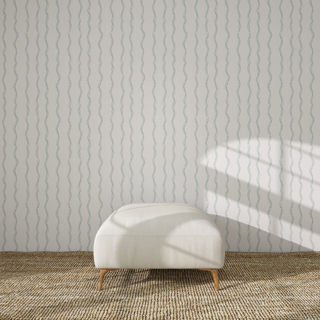A white upholstered bench sits on a woven rug in front of a light wall covered with Ribbon Stripe Wallpaper, Blue. Sunlight from the right casts geometric shadows across the wall.