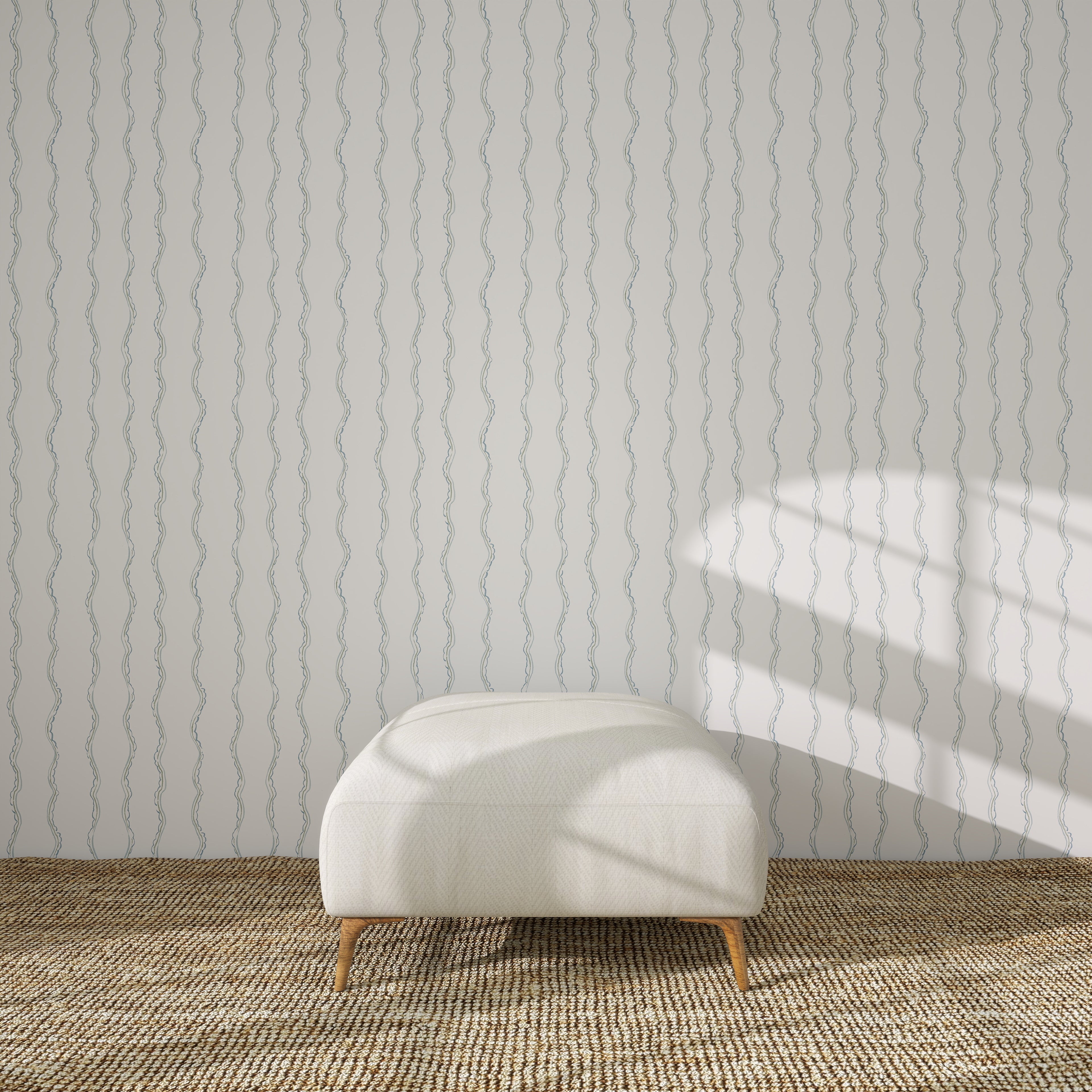 A white upholstered bench sits on a woven rug in front of a light wall covered with Ribbon Stripe Wallpaper, Blue. Sunlight from the right casts geometric shadows across the wall.