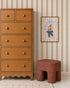 A wooden dresser with round knobs stands by a reddish-brown stool and framed art on a wall covered in Scallop Stripe Wallpaper, Pink. The light grid-patterned floor completes this playful kids’ room setup.