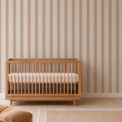 A wooden crib with a white mattress sits against a beige wall adorned with Scallop Stripe Wallpaper in Pink; a beige rug and part of a soft chair are visible in the nursery room.