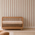 A wooden crib with a white mattress sits against a beige wall adorned with Scallop Stripe Wallpaper in Pink; a beige rug and part of a soft chair are visible in the nursery room.