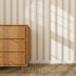 A wooden dresser stands on a woven rug beside a wall with the Scallop Stripe Wallpaper Sample, Pink. Sunlight streams in, casting diagonal shadows and highlighting the quality of the wallpaper.