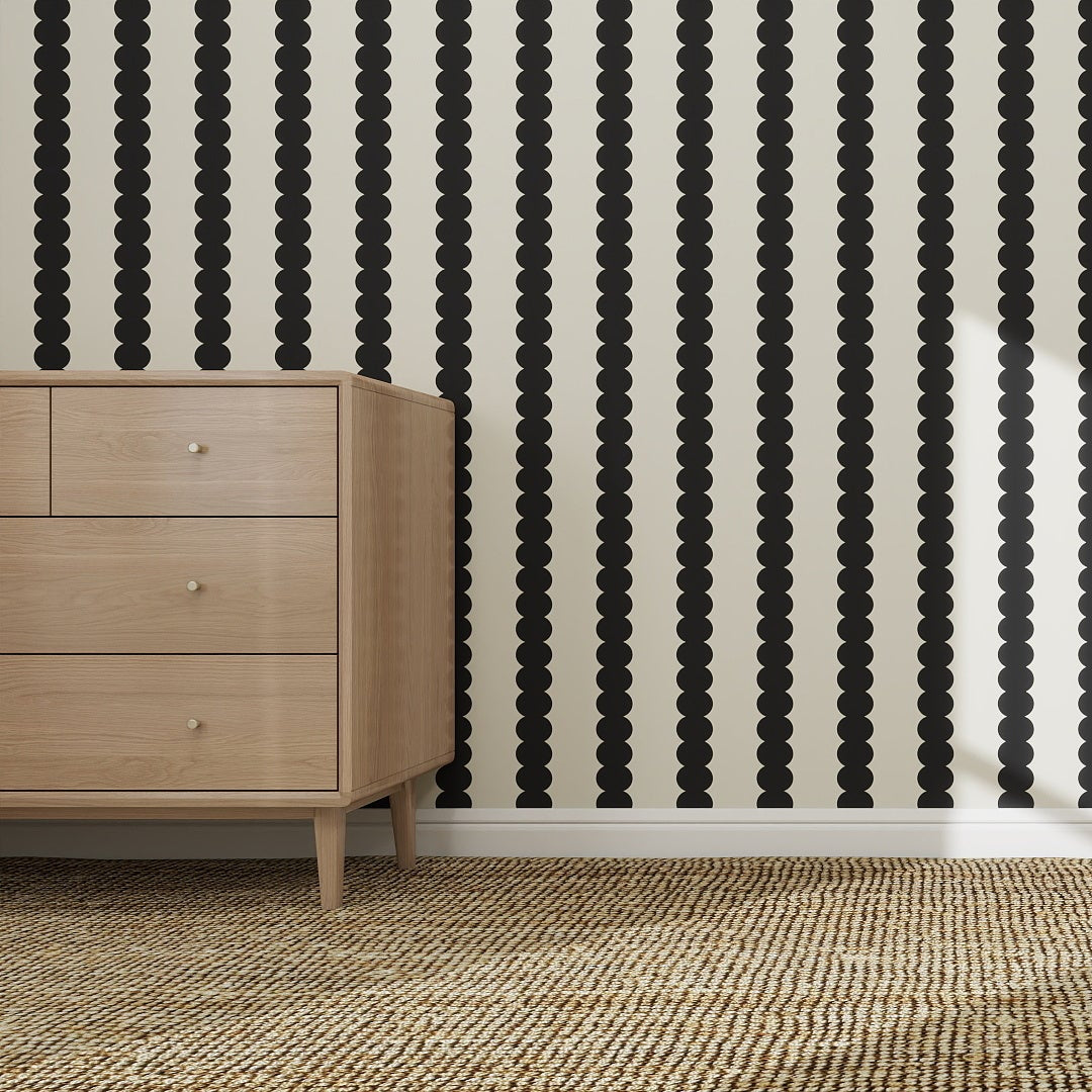 A light wood dresser with three drawers stands on a woven rug in front of Scallop Stripe Wallpaper in Charcoal, featuring vertical black polka dot stripes on a white background.