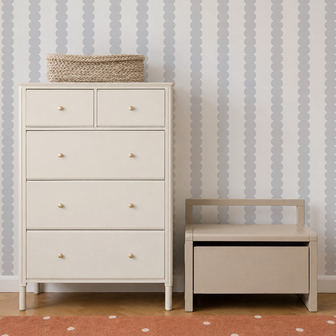 A cream-colored dresser with five drawers stands next to a small beige bench with a backrest, set against the charming Scallop Stripe Wallpaper in Grey. A woven basket rests atop the dresser, while an orange rug is partially visible beneath the soft blush pink accents in the room.