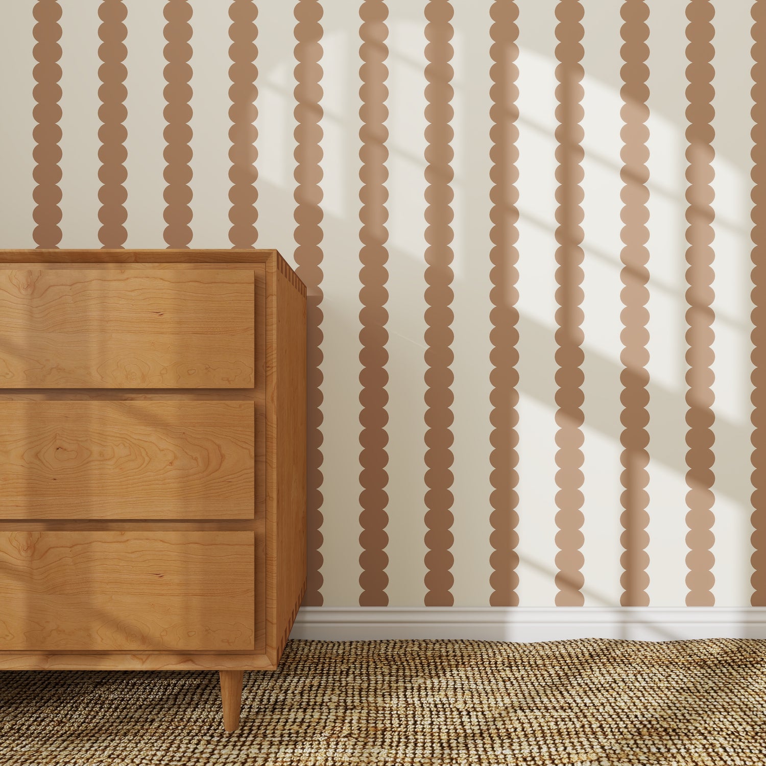A light wood dresser on a woven rug sits beside a wall decorated with Scallop Stripe Wallpaper, Latte. Sunlight casts diagonal shadows, bringing warmth and charm to this stylish nursery wallpaper setting.