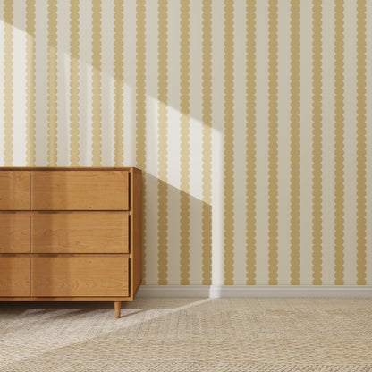 A wooden dresser with six drawers stands on a woven rug against a wall covered in Scallop Stripe Wallpaper Sample, Yellow. Sunlight streams in, casting diagonal shadows that accentuate the wallpaper’s unique yellow and beige stripes.