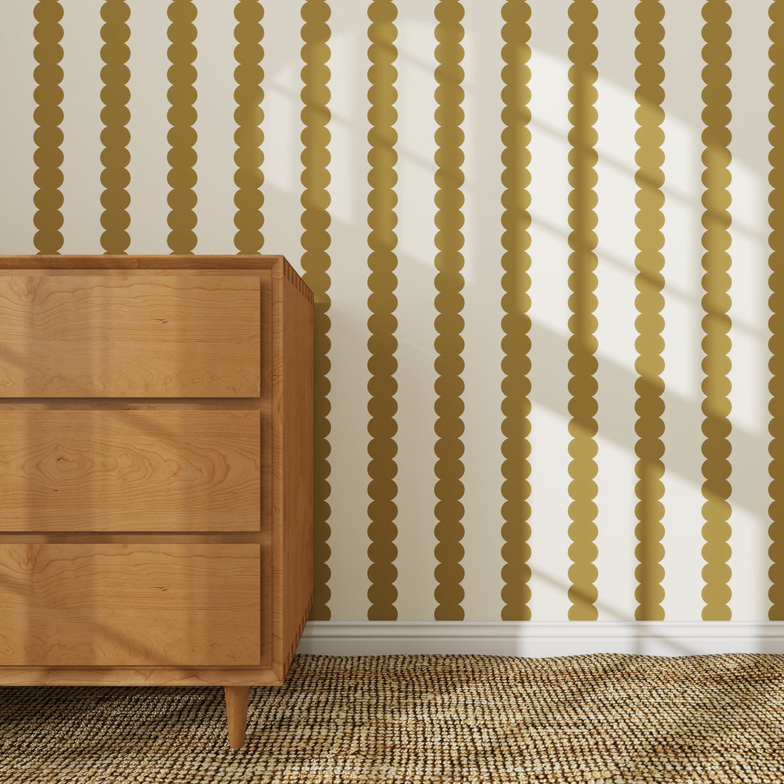 A wooden dresser stands on a woven rug beside a wall adorned with the Scallop Stripe Wallpaper Sample in Moss, its vertical gold circles catching the sunlight and casting soft shadows across the space.