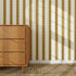 A wooden dresser stands on a woven rug beside a wall adorned with the Scallop Stripe Wallpaper Sample in Moss, its vertical gold circles catching the sunlight and casting soft shadows across the space.