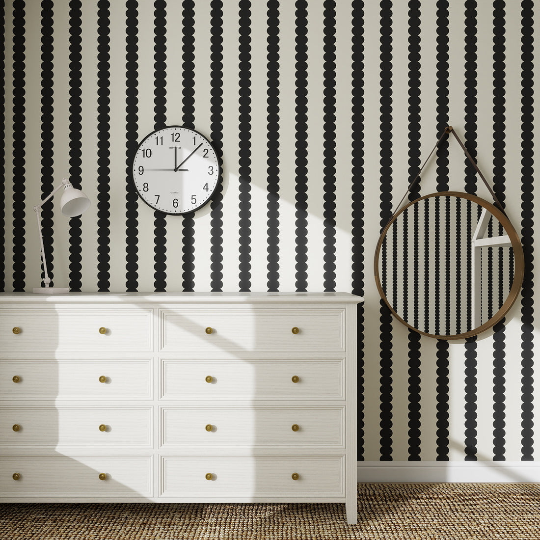 A white dresser with gold knobs stands against a wall covered in Scallop Stripe Wallpaper Charcoal. Above it hang a round clock and large round mirror with a brown strap, while a woven rug decorates the floor.