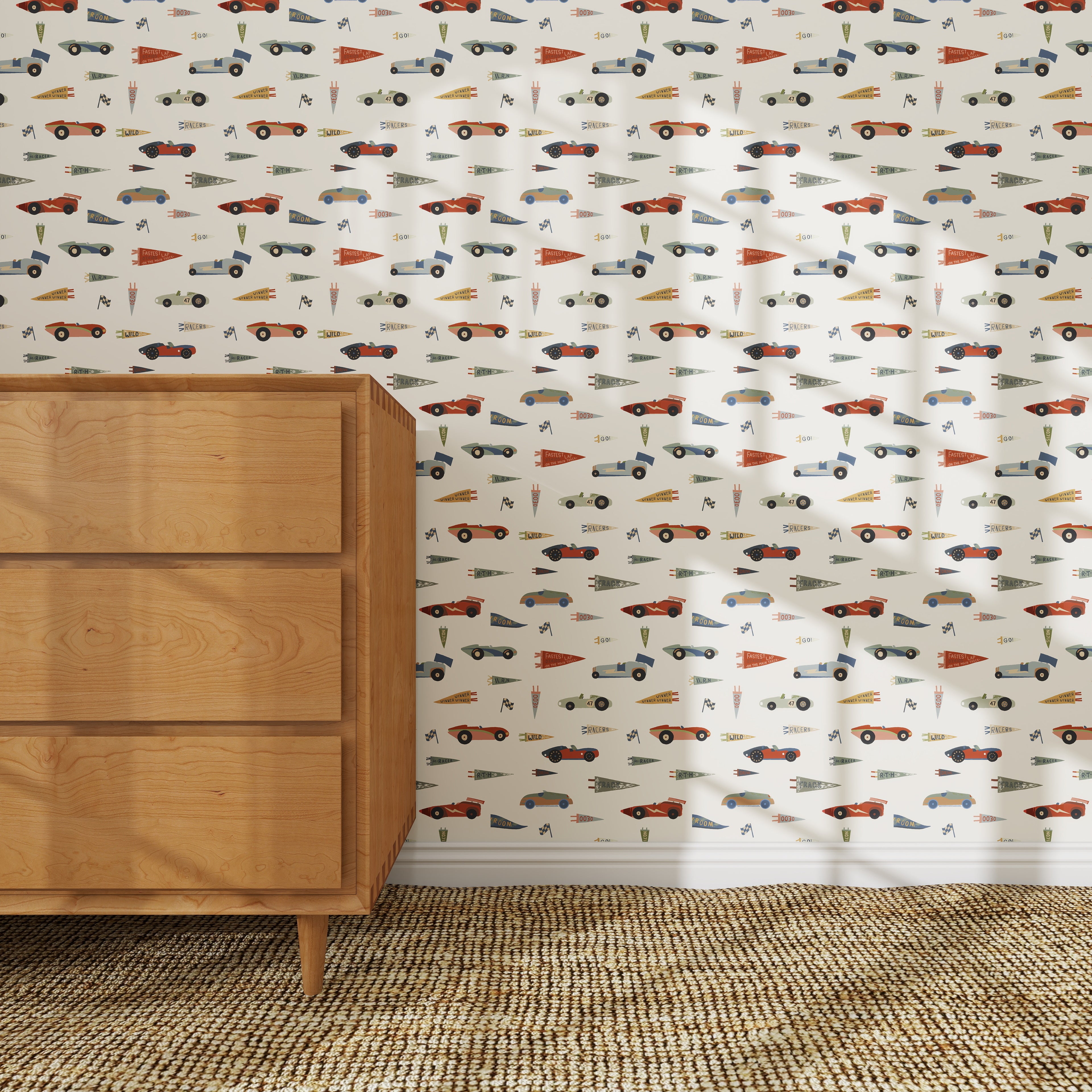 A light-filled room with a woven rug, a wooden mid-century dresser, and nursery wall decor featuring colorful vintage cars and pennants on Speedway Dash Wallpaper with a white background.