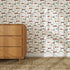 A light-filled room with a woven rug, a wooden mid-century dresser, and nursery wall decor featuring colorful vintage cars and pennants on Speedway Dash Wallpaper with a white background.