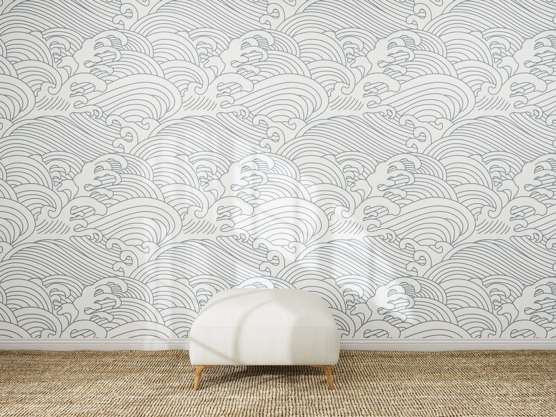 A white ottoman sits on a woven rug in front of a wall decorated with Making Waves Wallpaper, which displays a repeating pattern of hand-drawn gray ocean waves on a white background.