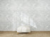 A white ottoman sits on a woven rug in front of a wall decorated with Making Waves Wallpaper, which displays a repeating pattern of hand-drawn gray ocean waves on a white background.