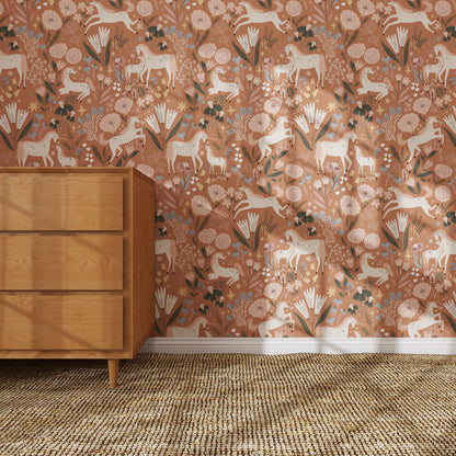 A wooden dresser stands on a woven rug by a wall covered in Unicorn Meadow Wallpaper Toffee Rose, featuring whimsical white deer, birds, flowers, and leaves in peach, pink, and green—perfect for charming girly room decor.