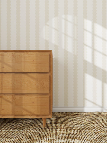 A wooden dresser with three drawers stands on a woven rug beside a wall adorned with Scallop Stripe Wallpaper Cloud, as sunlight streams in and casts window shadows across the wall and floor.