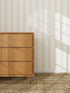 A wooden dresser with three drawers sits on a woven rug beside a wall adorned with Scallop Stripe Wallpaper in Cloud. Sunlight filters in, casting gentle shadows across the elegant scalloped pattern.