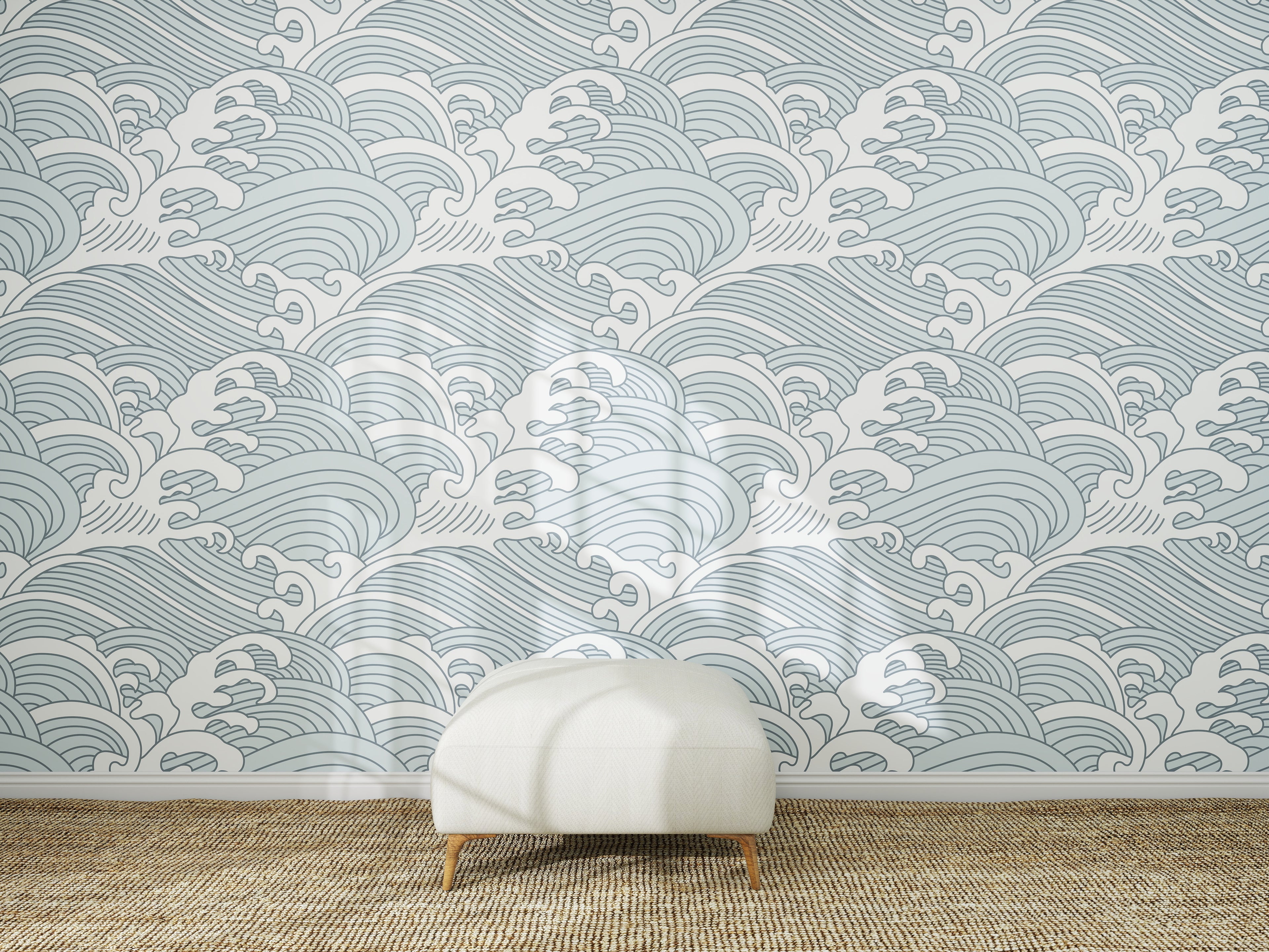 A white ottoman sits on a woven rug in front of a wall covered with Making Waves Wallpaper | Blue. Sunlight creates soft shadows on the floor and wallpaper mural.