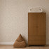 A brown bean bag sits next to a wooden wardrobe with two drawers, with a rolled-up blanket placed on top. The wall features Soaring Hearts Wallpaper, which has a light background and a small, subtle floral pattern.