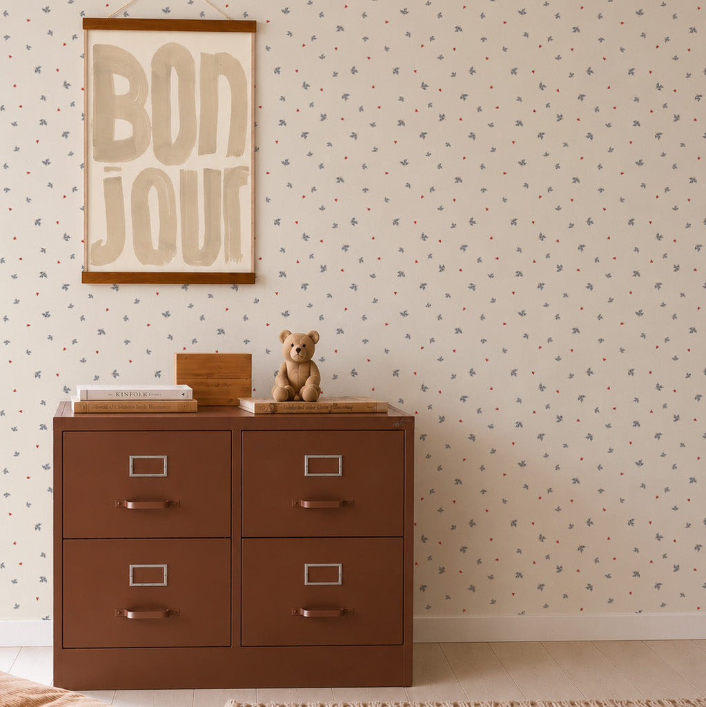 A brown four-drawer cabinet with a teddy bear and books on top sits against charming Soaring Hearts Wallpaper. Above, a framed BON JOUR print hangs on the wall, adding a delightful touch to the scene.
