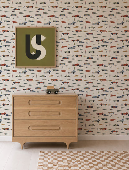 A wooden dresser stands against Speedway Dash Wallpaper, patterned with colorful vintage race cars and pennant flags on a cream background, topped with a small toy truck and a framed abstract print.