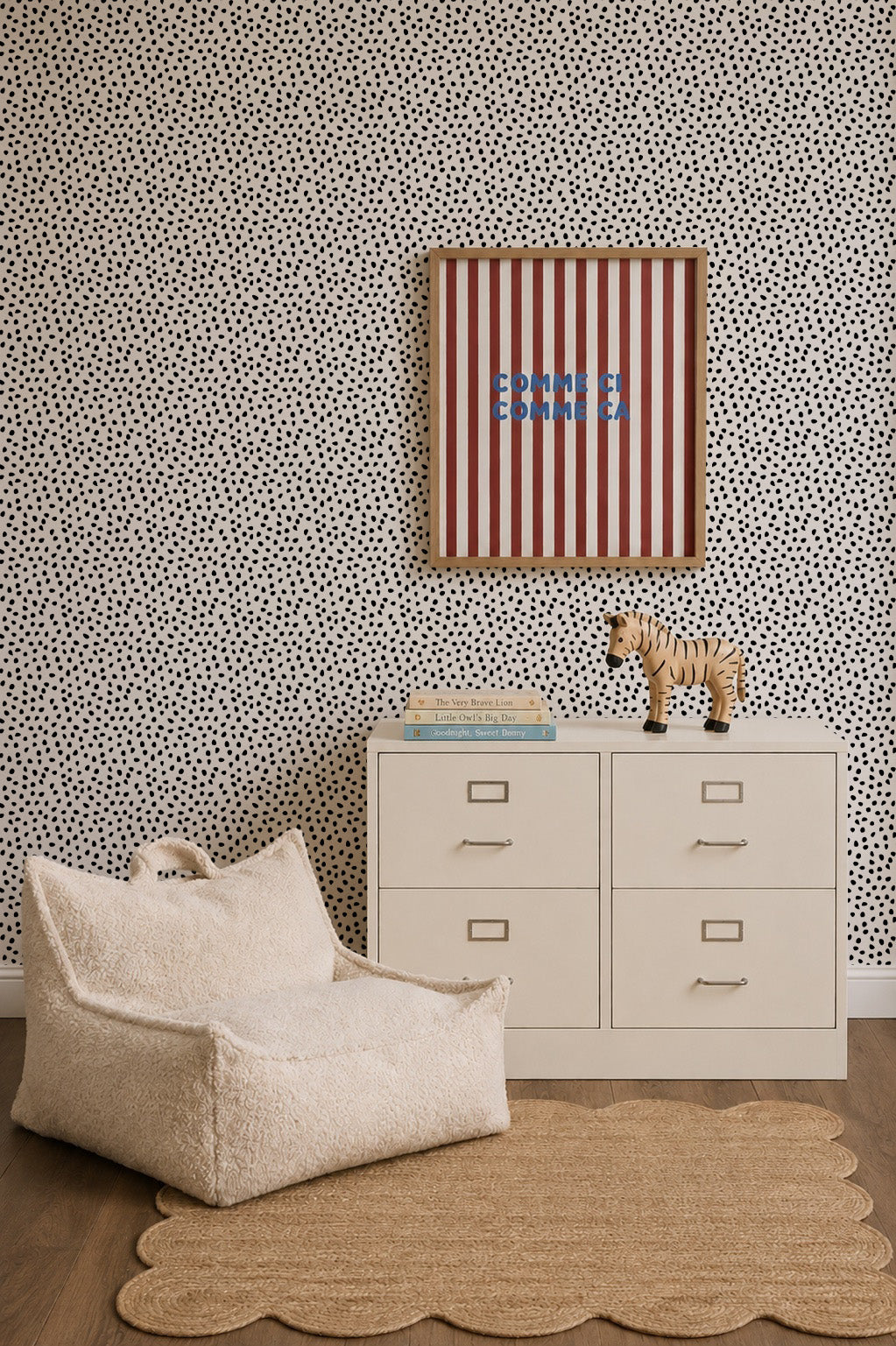 A modern nursery featuring Sprinkle Wallpaper in black, a white dresser, a plush chair, a scalloped beige rug, a toy zebra, books, and striped wall art with blue text.