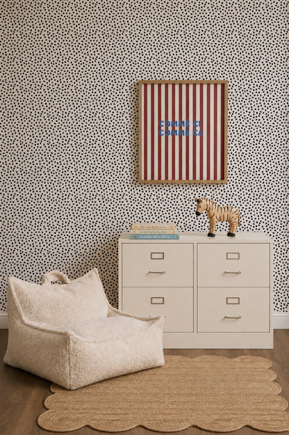 A modern nursery featuring Sprinkle Wallpaper in black, a white dresser, a plush chair, a scalloped beige rug, a toy zebra, books, and striped wall art with blue text.